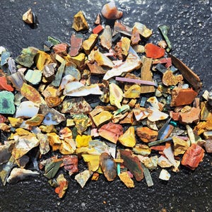 May include: A collection of colorful, irregularly shaped gemstones and minerals, including shades of red, yellow, green, and blue. The stones are scattered on a dark, textured surface, showcasing their varied textures and hues.