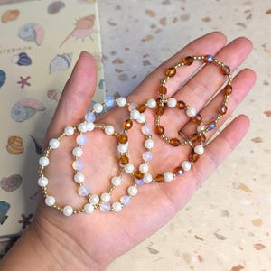 Pode incluir: Um conjunto de três pulseiras elásticas de contas com uma mistura de pérolas brancas, contas iridescentes translúcidas e pequenos espaçadores metálicos dourados. Uma pulseira destaca contas facetadas cor de âmbar e outras têm contas brancas.