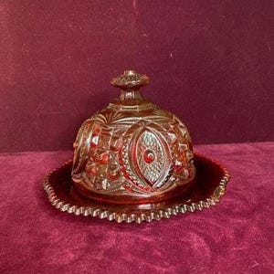 Puede incluir: Una mantequera antigua de cristal rojo rubí con tapa abovedada y borde decorativo festoneado. La tapa presenta un patrón detallado de cristal tallado con un diseño ovalado central. El plato se asienta sobre una placa a juego.