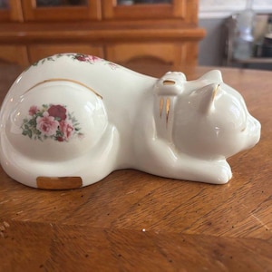 May include: A white ceramic cat figurine in a resting position, featuring a floral pattern of pink and red roses on its back and side. The cat wears a sculpted collar with gold trim, and there is a gold accent on its lower rear side.