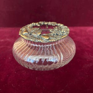 Puede incluir: Un florero de cristal transparente con cuerpo redondeado y acanalado, que incluye una tapa de latón extraíble. La tapa metálica presenta un patrón en relieve detallado de racimos de uvas y hojas de vid para sujetar los tallos.