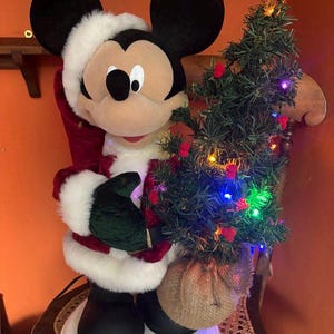 May include: A Mickey Mouse figurine dressed in a red and white Santa Claus suit with a matching hat, holding a small artificial Christmas tree decorated with colourful lights and red berries, standing on a round white base.