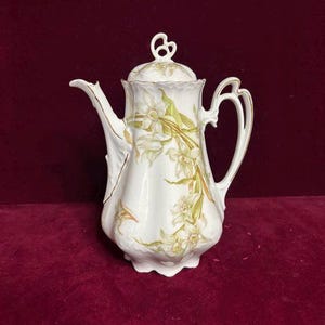 May include: White porcelain coffee pot with a lid, featuring a floral design in shades of green and yellow. The pot has a curved spout, a handle, and gold trim. A decorative piece for serving coffee or tea.