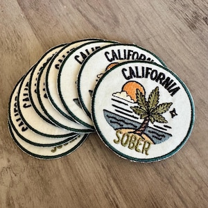 Peut inclure: Plusieurs écussons ronds brodés sur le thème de la Californie. Chaque écusson présente un palmier, un soleil et une scène océanique avec les mots "SOBER" et "CALIFORNIA" en fil noir et vert sur fond blanc.