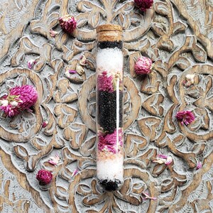 May include: A glass tube filled with bath salts, black sand, and dried pink flowers. The tube has a cork stopper and is sitting on a carved wooden surface.
