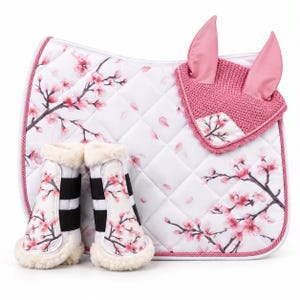 May include: A white quilted horse saddle pad, a pair of matching horse leg boots with cream faux fur lining, and a pink crocheted horse ear bonnet. All items feature a printed pattern of pink cherry blossom branches and scattered petals.