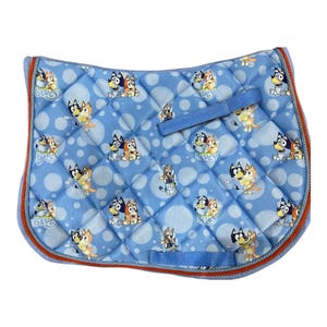 May include: A light blue quilted saddle pad featuring a repeating pattern of cartoon dogs. The pad has a blue border with orange trim and blue straps. The design includes white polka dots and the word "Bluey".