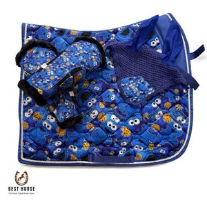 Könnte beinhalten: Ein blau gestepptes Reitsattelpad und passendes Zubehör mit einem sich wiederholenden Muster eines Zeichentrickcharakters und Keksen. Das Set enthält Gamaschen und eine Strickhülle. Die Kanten sind mit weißen Paspeln versehen.