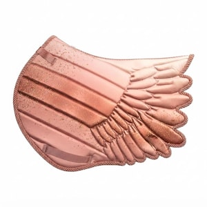 Puede incluir: Una mantilla de color oro rosa con un diseño acolchado en el lado izquierdo y una forma de ala tridimensional en el lado derecho. La superficie tiene un acabado brillante sutil y el borde está rematado con un cordón trenzado a juego.