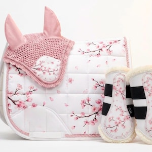 以下が含まれることがあります： ピンクの桜模様が施された白い馬用サドルパッド、お揃いのピンクのかぎ針編みイヤーネット、そして同じ花柄と黒いストラップ、縁にクリーム色の裏地が付いた白い馬用レッグブーツのセット。