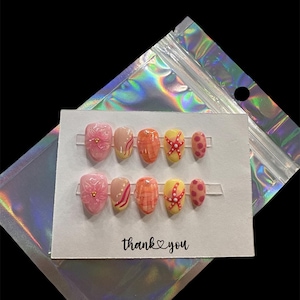 May include: A set of press-on nails with various designs, including pink floral, nude with yellow accents, orange plaid, yellow starfish, and pink polka dots. The card reads "thank you" in black script.