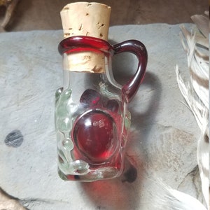 Może przedstawiać: Mała, przezroczysta butelka szklana z korkowym korkiem w kolorze czerwonym i czerwonym szklanym uchwytem. Butelka jest wypełniona czerwonym płynem i ma fakturowaną powierzchnię.