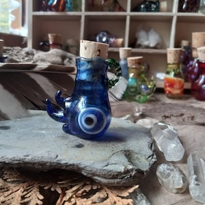 May include: A small blue glass bottle with a cork stopper and a blue evil eye charm. The bottle is sitting on a gray stone.