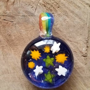May include: A round glass pendant with a rainbow-colored top and a blue background. The pendant features white stars, yellow suns, and green leaves.
