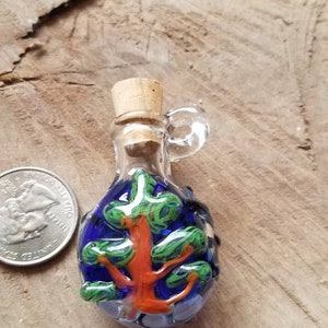 May include: A small glass bottle with a cork stopper and a loop handle. The bottle is decorated with a blue background and a colorful tree design.