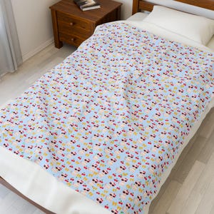 May include: A light blue blanket featuring a repeating pattern of cartoon cats wearing sunglasses, cherries, and yellow bows. The blanket is on a bed with a white pillow and a wooden bedside table.