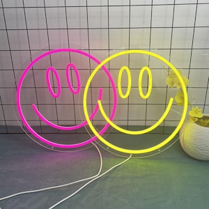 Könnte beinhalten: Zwei Neon-Smiley-Gesichter. Eines ist leuchtend pink, das andere gelb. Beide sind kreisförmig mit transparentem Hintergrund und weißen Netzkabeln. Die Schilder stehen auf einer grauen Oberfläche vor einer weißen Fliesenwand.