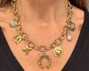 May include: A gold-coloured charm necklace with a chunky chain and various pendants. Charms include a horseshoe, star, heart, and geometric shapes. The necklace has a mix of textures and details. A statement piece of jewellery.