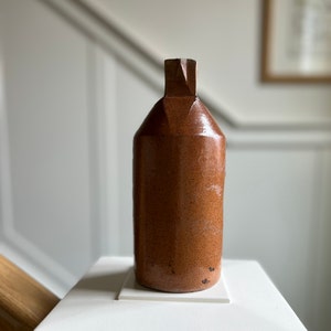 May include: A cylindrical brown stoneware bottle with a glossy, speckled finish and a narrow, squared-off neck. The vessel features a uniform, earthy tone with subtle variations in texture across its surface, standing upright with a flat base.
