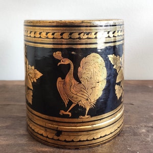 May include: Black cylindrical container with gold accents and a peacock design. The container has a decorative pattern around the top and bottom.