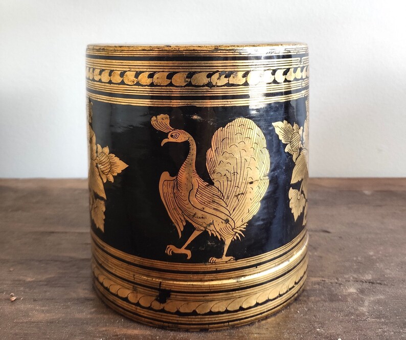 May include: Black cylindrical container with gold accents and a peacock design. The container has a decorative pattern around the top and bottom.