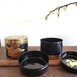 May include: Three black and gold cylindrical boxes with intricate designs. The boxes are made of lacquered wood and feature gold leaf accents. One box has a floral design, another has a horse design, and the third is plain.