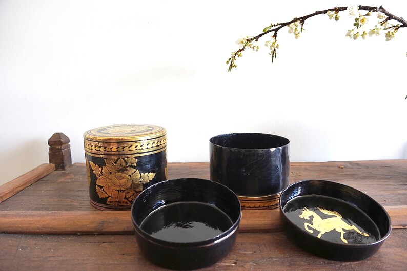 May include: Three black and gold cylindrical boxes with intricate designs. The boxes are made of lacquered wood and feature gold leaf accents. One box has a floral design, another has a horse design, and the third is plain.