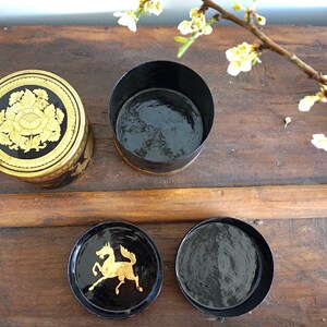 May include: Four round black boxes with gold accents. The boxes have different designs on the top. One box has a floral design, another has a horse design, and the other two are plain.