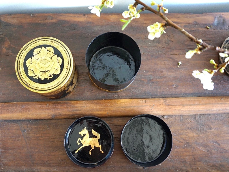 May include: Four round black boxes with gold accents. The boxes have different designs on the top. One box has a floral design, another has a horse design, and the other two are plain.