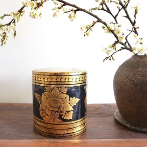 May include: A black and gold cylindrical box with intricate floral designs. The box is decorated with gold trim and has a traditional Asian aesthetic.