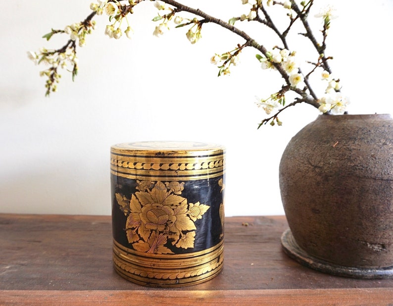 May include: A black and gold cylindrical box with intricate floral designs. The box is decorated with gold trim and has a traditional Asian aesthetic.