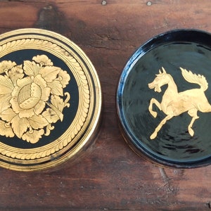 May include: Two round, black lacquered boxes with gold painted designs. One box features a floral design, while the other has a gold horse design.