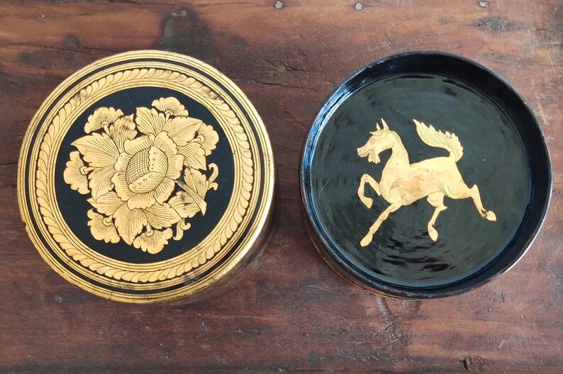 May include: Two round, black lacquered boxes with gold painted designs. One box features a floral design, while the other has a gold horse design.