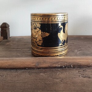 May include: A black cylindrical box with gold accents and intricate designs. The box features a peacock and floral motifs.