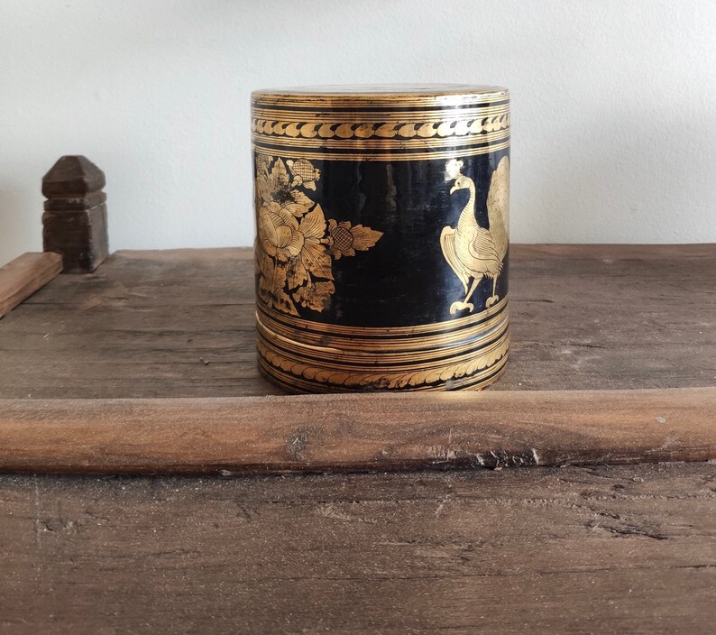 May include: A black cylindrical box with gold accents and intricate designs. The box features a peacock and floral motifs.