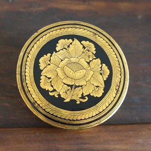 May include: A round, black and gold decorative trinket box with a floral design. The box is made of wood and has a gold rim.