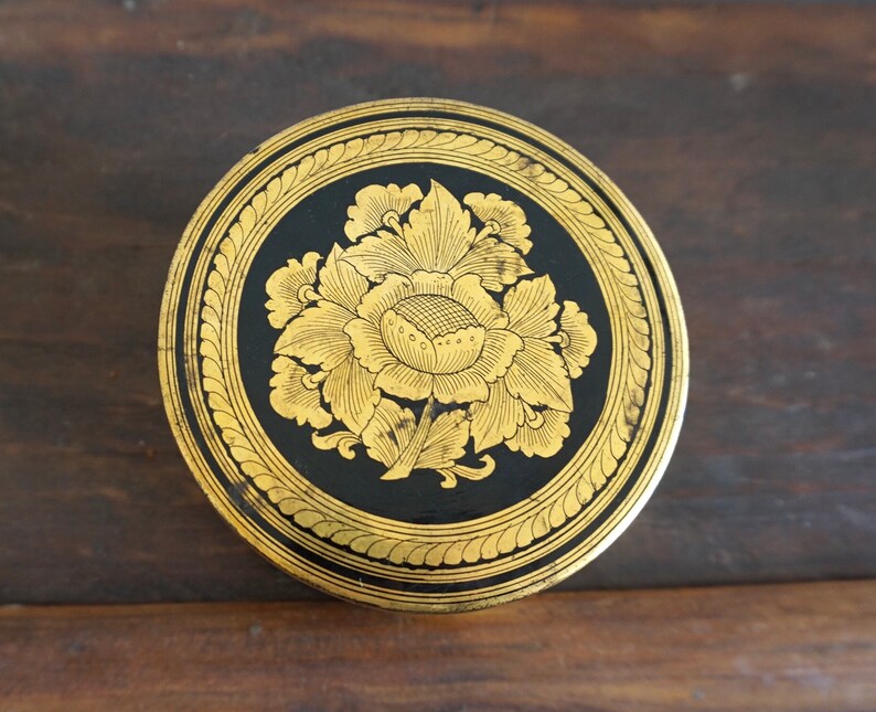 May include: A round, black and gold decorative trinket box with a floral design. The box is made of wood and has a gold rim.