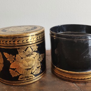 May include: Two black cylindrical boxes with gold floral designs. The boxes are made of lacquered wood and have a shiny finish.
