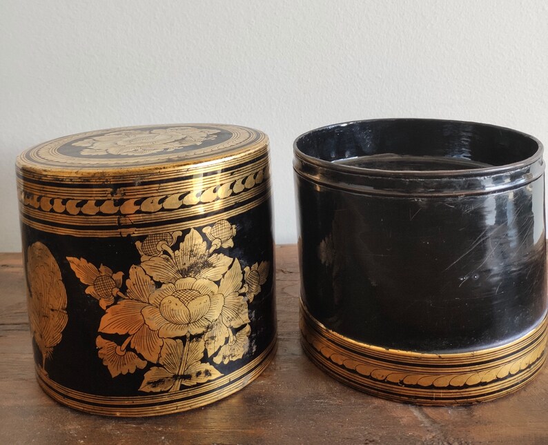 May include: Two black cylindrical boxes with gold floral designs. The boxes are made of lacquered wood and have a shiny finish.