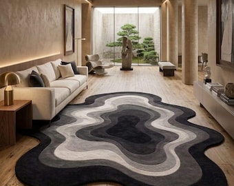 May include: A modern living room featuring a large, abstract-shaped rug in shades of gray and black. The rug sits on a wooden floor, with a white sofa and a view of a serene outdoor garden through a large window.