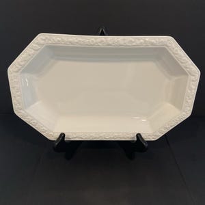 May include: A white ceramic serving platter with an elongated octagonal shape. The rim features a raised, embossed floral and leaf pattern that runs along the entire perimeter of the dish. The center of the platter is smooth, glossy, and flat.