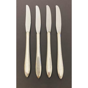 Könnte beinhalten: Vier Buttermesser aus Edelstahl mit reflektierender, silberner Oberfläche. Die Messer haben ein schlichtes, elegantes Design mit gebogener Klinge und dekorativem Griff. Die Messer sind vertikal vor einem dunklen Hintergrund angeordnet.