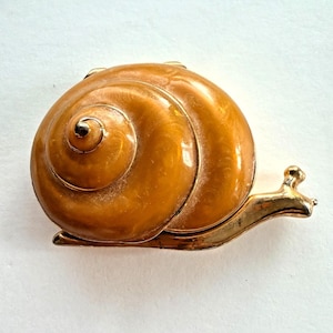 May include: A snail-shaped brooch featuring a glossy, amber-colored enamel shell with a swirling pattern. The body of the snail is crafted from a metallic gold-toned material, extending from beneath the shell to form the head and tail sections.