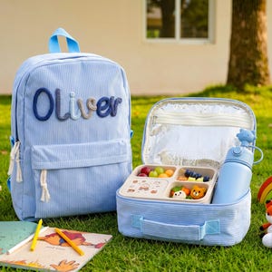 Puede incluir: Una mochila de pana azul claro con el nombre Oliver bordado en hilo azul oscuro y beige, junto a una bolsa térmica a juego que contiene un recipiente de comida dividido y una botella de agua azul claro con una tapa en forma de gato.