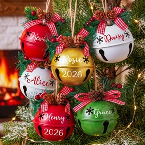 Puede incluir: Un conjunto de cinco adornos navide&ntilde;os de metal en forma de cascabel redondo en colores rojo, blanco, dorado y verde. Cada uno lleva un nombre personalizado, algunos incluyen el a&ntilde;o 2026 y est&aacute;n decorados con pi&ntilde;as y lazos de cuadros rojos.