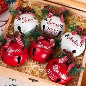 May include: A set of six round metal jingle bell ornaments in red and white, each personalized with names like Oliver, Lucas, Grace, Daniel, Amelia, and Clara. Some feature the year 2026, topped with pine needles, pinecones, and red plaid ribbons.