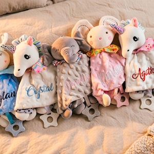 Puede incluir: Una colección de mantas de seguridad para bebés con cabezas de animales, incluyendo un conejito, un unicornio y un elefante. Cada manta es de un color diferente y tiene un nombre bordado. Las mantas tienen mordedores en forma de estrella.