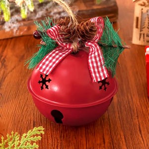 Puede incluir: Un adorno de cascabel de metal rojo con recortes en forma de copo de nieve, decorado con un lazo de cuadros rojos y blancos, pi&ntilde;as peque&ntilde;as, ramas de pino artificiales y un cordel de yute para colgar en el &aacute;rbol.