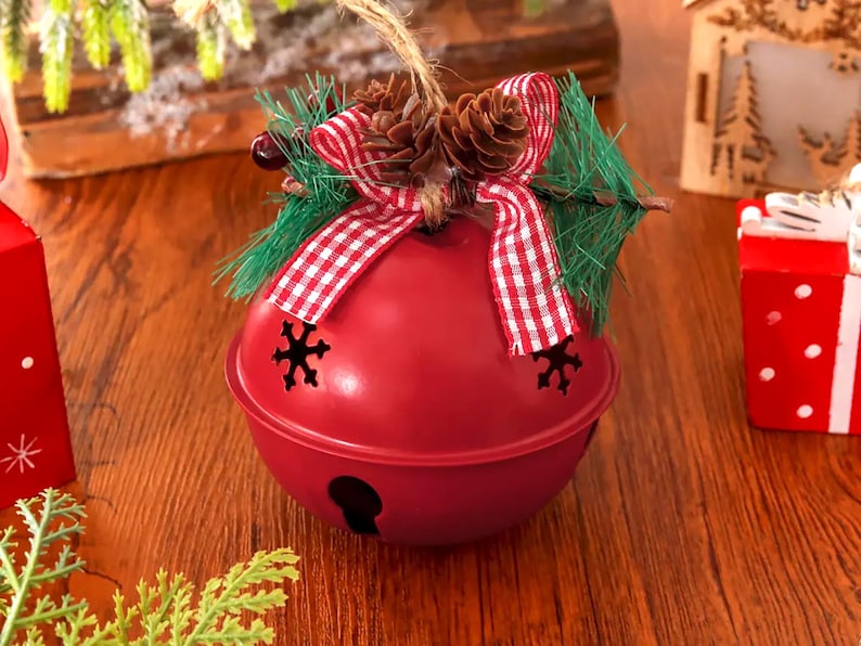 Puede incluir: Un adorno de cascabel de metal rojo con recortes en forma de copo de nieve, decorado con un lazo de cuadros rojos y blancos, pi&ntilde;as peque&ntilde;as, ramas de pino artificiales y un cordel de yute para colgar en el &aacute;rbol.