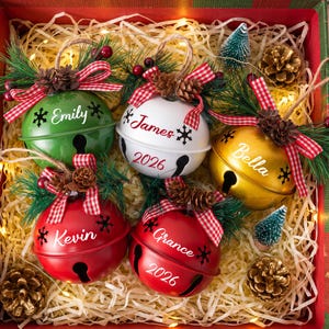 Puede incluir: Cinco adornos de cascabeles met&aacute;licos redondos en verde, blanco, dorado y rojo, personalizados con nombres como Emily, James, Bella, Kevin y Grance, decorados con lazos de cuadros rojos, agujas de pino y pi&ntilde;as peque&ntilde;as.